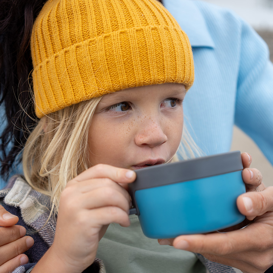 Ein Kind mit einer gelben Mütze sitzt bei seiner Mutter und trinkt einen Tee.
