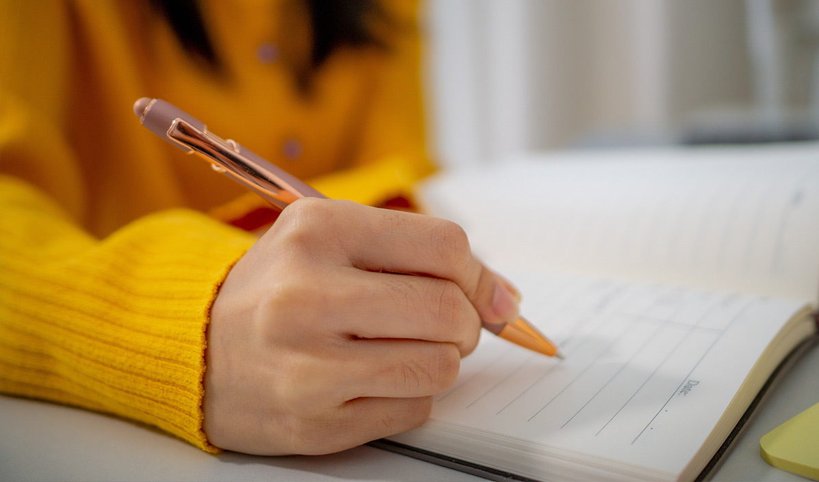 Eine Frau in einem gelben Pullover sitzt am Schreibtisch und schreibt etwas auf.
