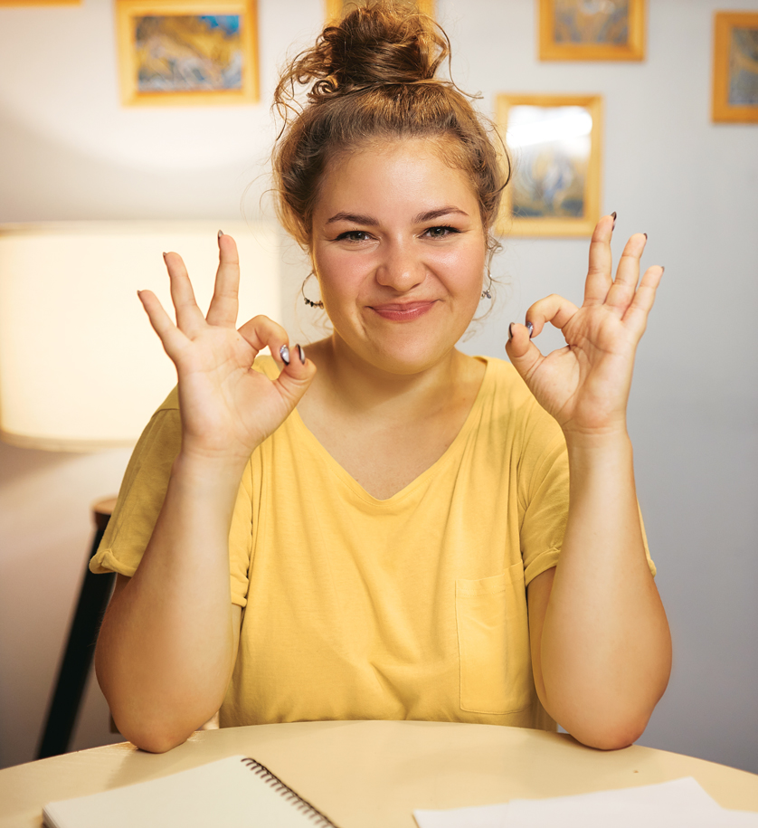 Eine Frau mit gelbem T-Shirt sitzt an einem Tisch, lächelt und hat die Hände zu einem "Super-Zeichen" geformt.